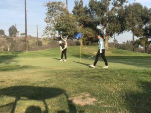 Two golfers on the green, with one preparing to putt and the other observing, illustrating the concept of golf handicap and slope, with trees and hills in the background.